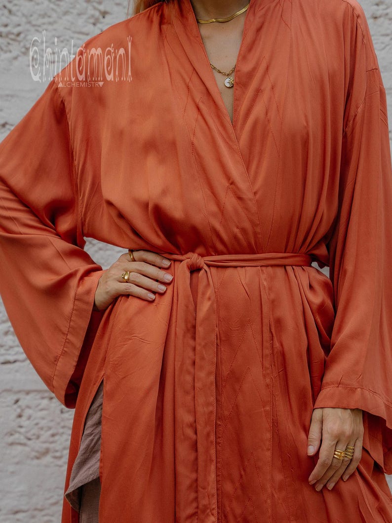 May include: A burnt orange, long-sleeved robe with a wrap-around design and a matching belt. The robe has a silky texture and features subtle vertical stitching details. The sleeves are wide and flowing. The brand name "chintamani" is visible.