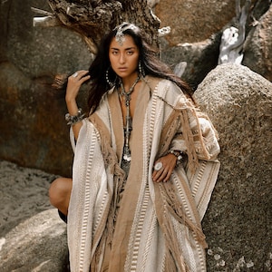 Puede incluir: Un poncho tejido en color beige y crema con rayas verticales texturizadas y flecos largos en el dobladillo. La prenda tiene un dise&ntilde;o abierto en la parte delantera con un panel central marr&oacute;n claro y detalles trenzados complejos.