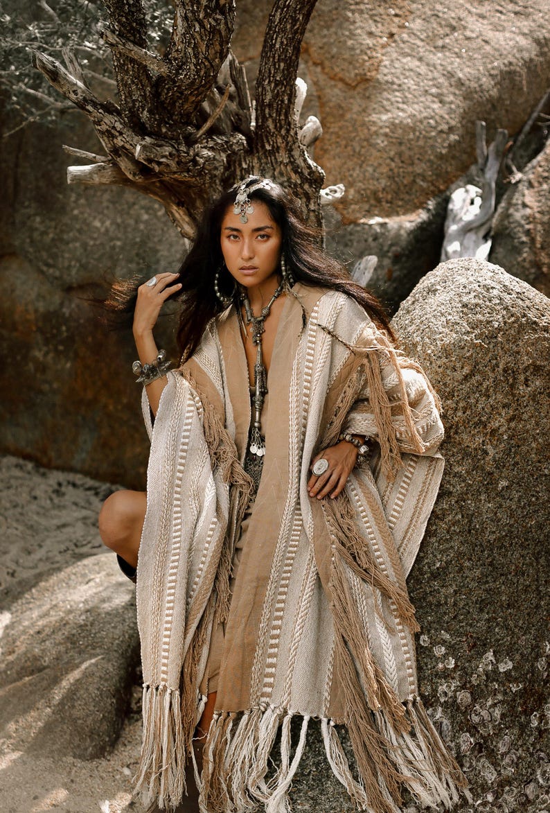 Puede incluir: Un poncho tejido en color beige y crema con rayas verticales texturizadas y flecos largos en el dobladillo. La prenda tiene un dise&ntilde;o abierto en la parte delantera con un panel central marr&oacute;n claro y detalles trenzados complejos.