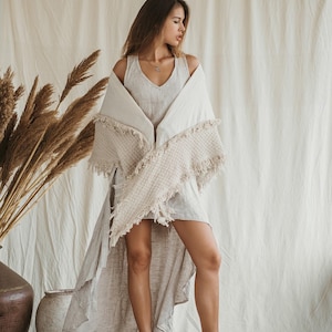 Puede incluir: Un vestido beige sin mangas con escote en V y dobladillo asimétrico, combinado con un chal color crema texturizado con bordes deshilachados. El conjunto se completa con botines claros que tienen múltiples correas con hebillas.