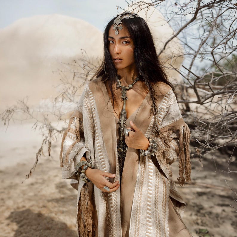 Puede incluir: Un poncho con estampado en beige y crema con detalles de flecos largos, usado con un tocado plateado, un collar largo con colgante plateado, m&uacute;ltiples pulseras plateadas y anillos de plata. La prenda presenta un tejido texturizado y escote en V.