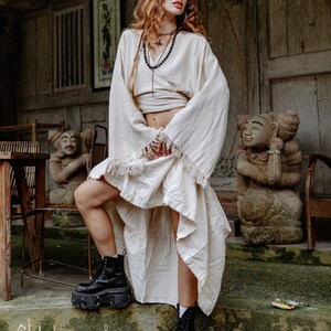 Puede incluir: Conjunto crema de top envolvente de manga larga y falda con flecos. La modelo lleva botas de plataforma negras. La imagen incluye el texto "chintamani alchemy" en la parte inferior.