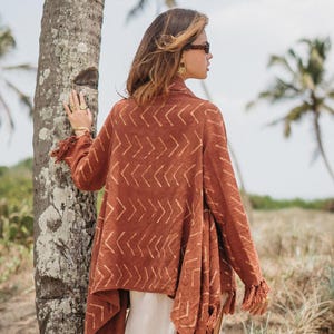 May include: A terracotta-coloured open cardigan with a geometric arrow pattern and fringing. The cardigan is worn over a white outfit. The image includes the text "Chintamani" in the upper right corner.