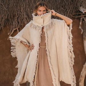 May include: A beige textured poncho featuring a high, structured collar with snap buttons and frayed edges along the hem and front opening. The garment is worn over a solid brown dress. The text Chintamani Alchemistry is visible in the top corner.