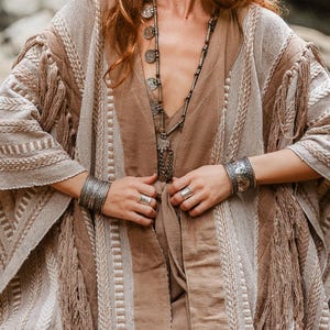 Puede incluir: Un kimono texturizado en beige y marr&oacute;n claro con flecos, usado sobre una prenda de color marr&oacute;n claro s&oacute;lido. El conjunto se complementa con un collar largo plateado con colgantes circulares, dos brazaletes anchos de metal plateado y dos anillos plateados.