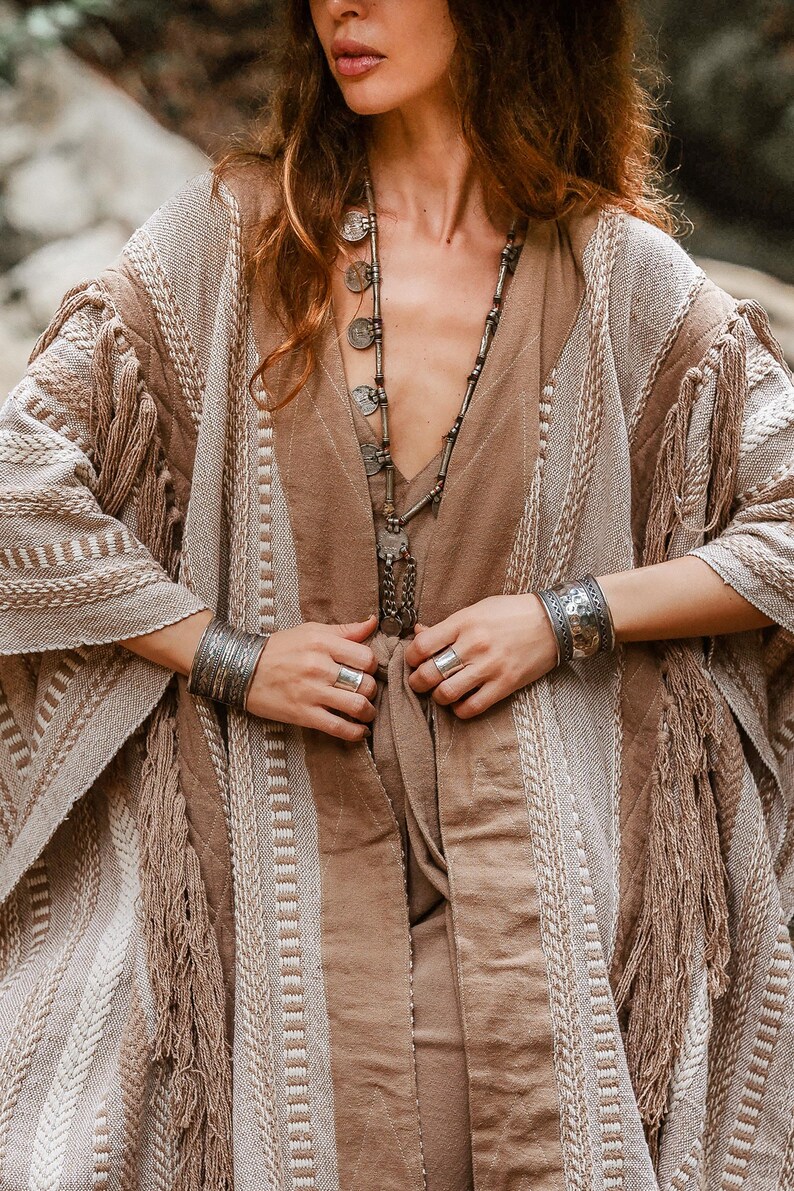Puede incluir: Un kimono texturizado en beige y marr&oacute;n claro con flecos, usado sobre una prenda de color marr&oacute;n claro s&oacute;lido. El conjunto se complementa con un collar largo plateado con colgantes circulares, dos brazaletes anchos de metal plateado y dos anillos plateados.