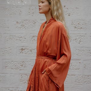 May include: An orange-red robe with wide sleeves and a matching belt. The robe has a relaxed fit and a pocket on the side. The word "Chintamani" is printed in white on the top left corner. The robe is made of a silky material.