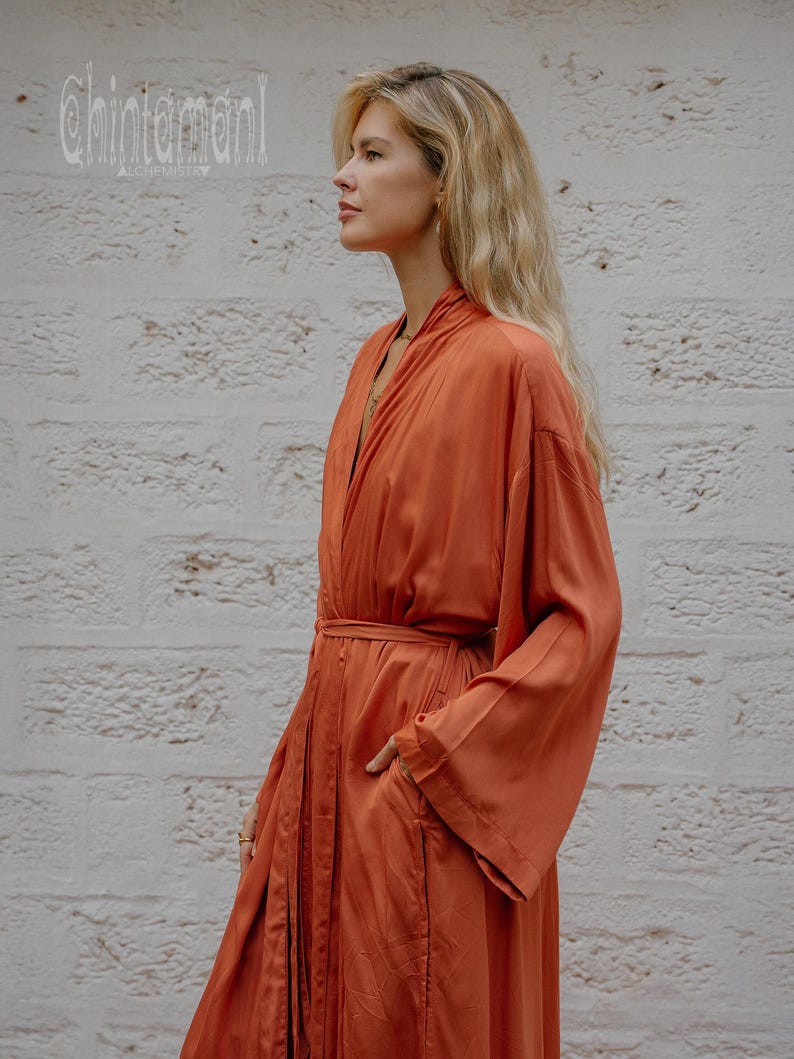 May include: An orange-red robe with wide sleeves and a matching belt. The robe has a relaxed fit and a pocket on the side. The word "Chintamani" is printed in white on the top left corner. The robe is made of a silky material.