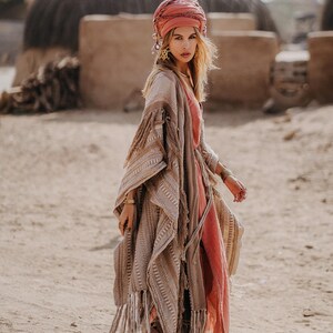 Puede incluir: Un poncho beige con estampado y bordes de flecos largos sobre un vestido color coral. El conjunto incluye un pa&ntilde;uelo para la cabeza color coral, joyas doradas y botas marrones. El poncho presenta texturas tejidas intrincadas y una silueta.