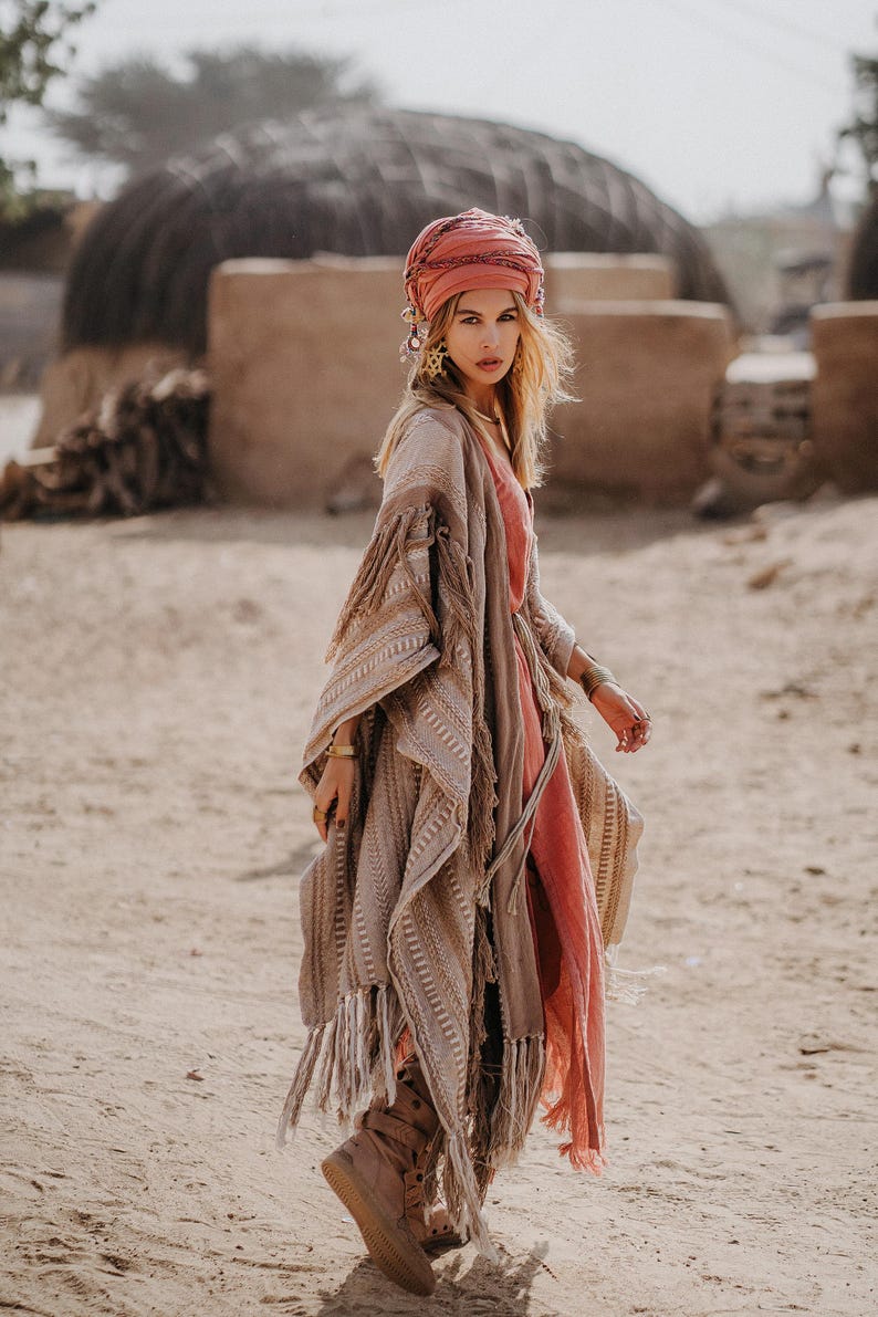 Puede incluir: Un poncho beige con estampado y bordes de flecos largos sobre un vestido color coral. El conjunto incluye un pa&ntilde;uelo para la cabeza color coral, joyas doradas y botas marrones. El poncho presenta texturas tejidas intrincadas y una silueta.