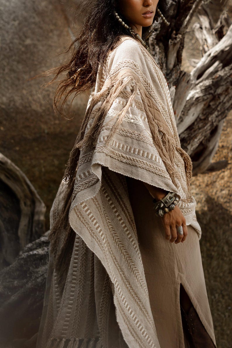 Puede incluir: Un poncho de color beige con intrincadas texturas tejidas, patrones trenzados y detalles de flecos en los bordes. La prenda se superpone sobre un vestido beige liso con una abertura lateral, complementado con un brazalete y un anillo.