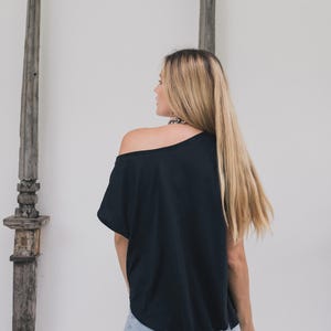 May include: Black off-the-shoulder top paired with light blue jeans. The model is facing away from the camera, showcasing the back of the top and jeans. The logo "chintamani alchemy" is visible at the bottom left.