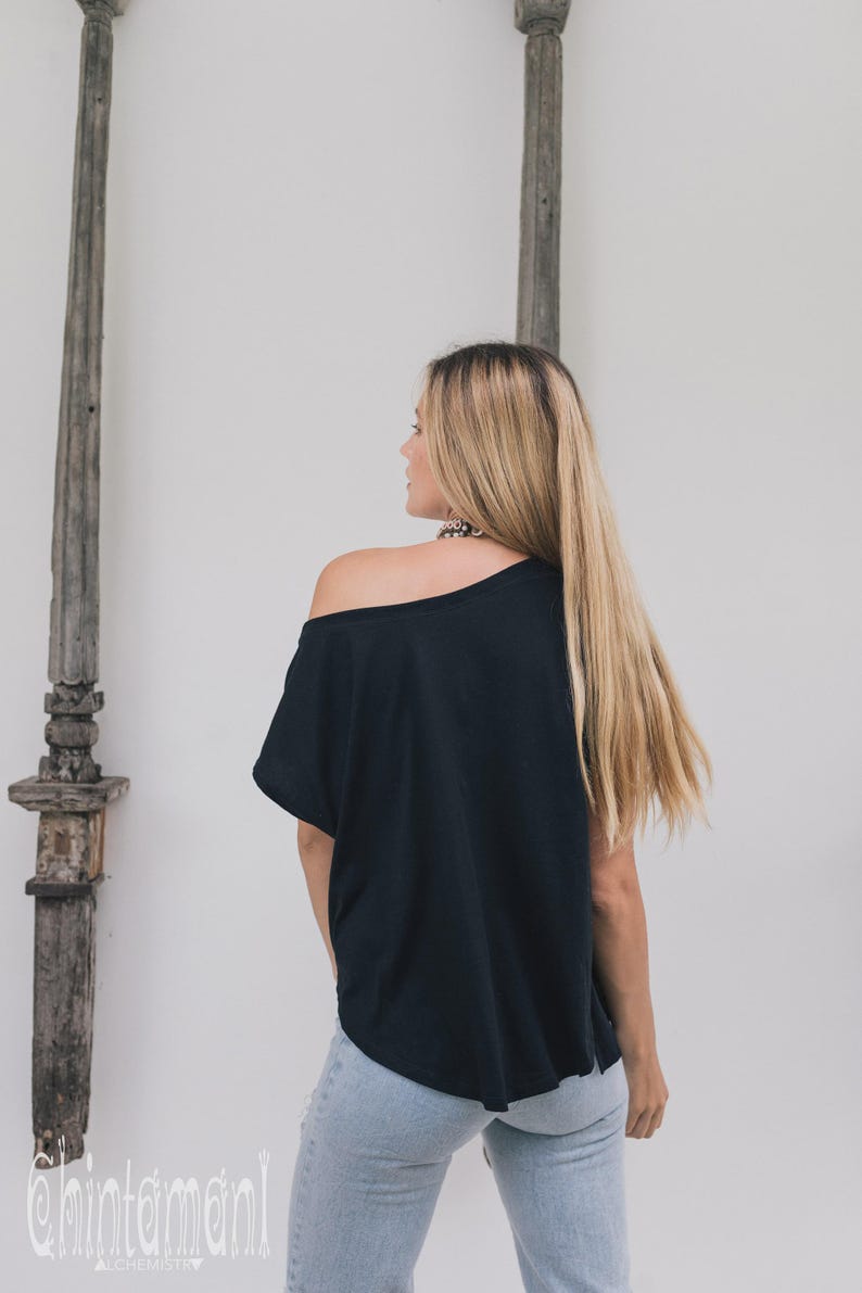 May include: Black off-the-shoulder top paired with light blue jeans. The model is facing away from the camera, showcasing the back of the top and jeans. The logo "chintamani alchemy" is visible at the bottom left.
