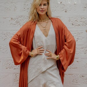 May include: A woman in a rust-coloured kimono-style robe over a light grey jumpsuit. The outfit is accessorised with gold jewellery, including necklaces, bracelets, and rings. The background is a white brick wall. The word "Chintamani" is visible in the upper right corner.