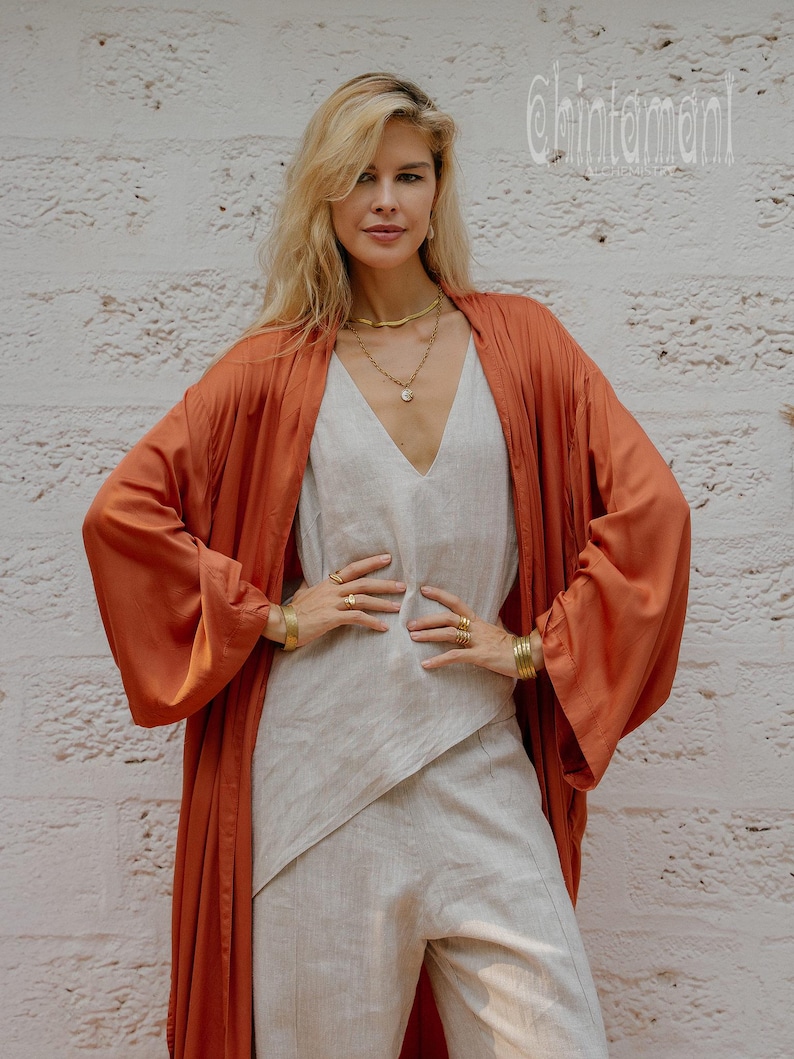 May include: A woman in a rust-coloured kimono-style robe over a light grey jumpsuit. The outfit is accessorised with gold jewellery, including necklaces, bracelets, and rings. The background is a white brick wall. The word "Chintamani" is visible in the upper right corner.