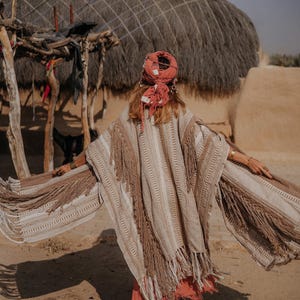 Puede incluir: Un poncho de color beige y tostado con rayas verticales texturizadas y flecos largos en los bordes. La prenda se lleva con un pa&ntilde;uelo de color coral y una falda a juego, combinada con botas acordonadas de color beige y pulseras doradas.