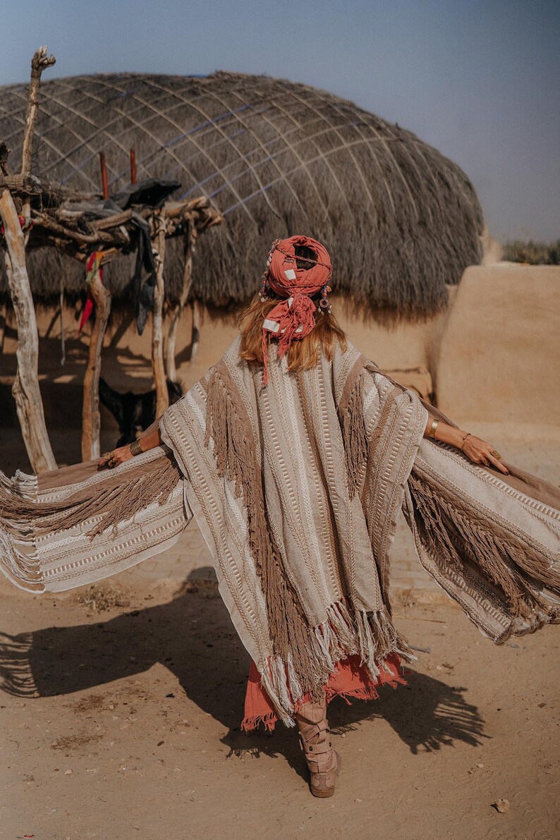 Puede incluir: Un poncho de color beige y tostado con rayas verticales texturizadas y flecos largos en los bordes. La prenda se lleva con un pa&ntilde;uelo de color coral y una falda a juego, combinada con botas acordonadas de color beige y pulseras doradas.