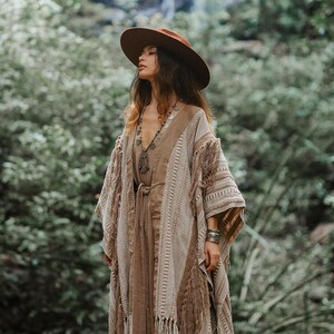 Puede incluir: Un sombrero marr&oacute;n de ala ancha, un vestido sin mangas color canela con lazo en la cintura y un kimono abierto texturizado en beige y marr&oacute;n con patrones de punto verticales y flecos largos en los bordes, junto a un collar con colgante y pulsera.