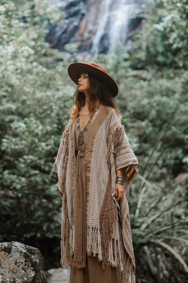 Puede incluir: Un sombrero marr&oacute;n de ala ancha, un vestido sin mangas color canela con lazo en la cintura y un kimono abierto texturizado en beige y marr&oacute;n con patrones de punto verticales y flecos largos en los bordes, junto a un collar con colgante y pulsera.
