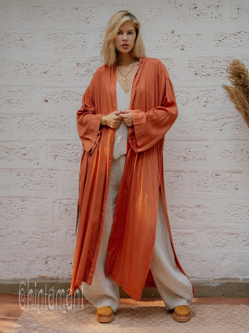 May include: A long, flowing rust-coloured robe with wide sleeves and a high slit, worn over a white top and wide-leg trousers. The robe has a silky sheen. The model is wearing gold jewellery. The brand name "chintamani" is visible at the bottom.