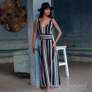 Può includere: Un abito lungo nero senza maniche con scollo a V e un motivo geometrico bianco. L'abito include una cintura da annodare abbinata e una gonna lunga e fluida. Un cappello nero a tesa larga è indossato con l'abito dall'orlo sfrangiato.