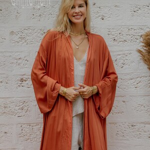 May include: A woman in a rust-coloured, open-front robe over a light-coloured outfit. The robe has long sleeves and a flowing design. The background is a textured, white brick wall. The text "chintamani" is visible in the upper left corner.