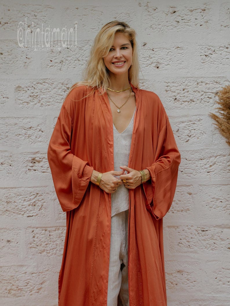 May include: A woman in a rust-coloured, open-front robe over a light-coloured outfit. The robe has long sleeves and a flowing design. The background is a textured, white brick wall. The text "chintamani" is visible in the upper left corner.