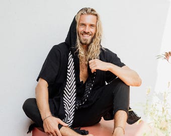 May include: A black hooded short-sleeve tunic featuring a white chevron-patterned trim along the front opening. The garment has a relaxed fit and includes the text Chintamani Alchemistry printed in white in the upper left corner of the image.