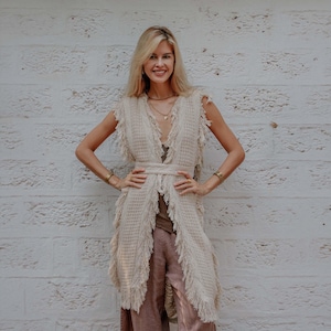 May include: A sleeveless, beige, open-front waistcoat featuring a textured knit pattern and long fringe detailing along the edges. The garment includes a matching tie-waist belt and is worn over a dark brown top and loose-fitting brown trousers.