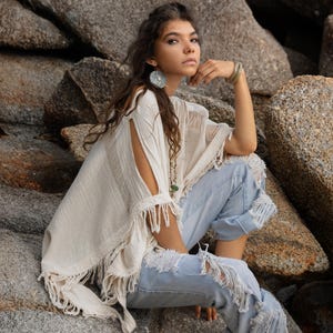Könnte beinhalten: Eine Frau trägt einen cremefarbenen Poncho mit Fransen und eine zerrissene hellblaue Jeans. Der Poncho hat offene Ärmel und ein fließendes Design. Die Jeans hat gerissene Details. Der Hintergrund zeigt große Felsen und den Markennamen "Chintamani Alchemy".