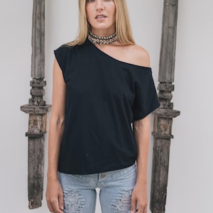 May include: Black off-the-shoulder top paired with light blue distressed jeans. The top has a single short sleeve and a unique asymmetrical neckline. A beaded choker necklace adds a touch of detail. The background features wooden decorative elements.