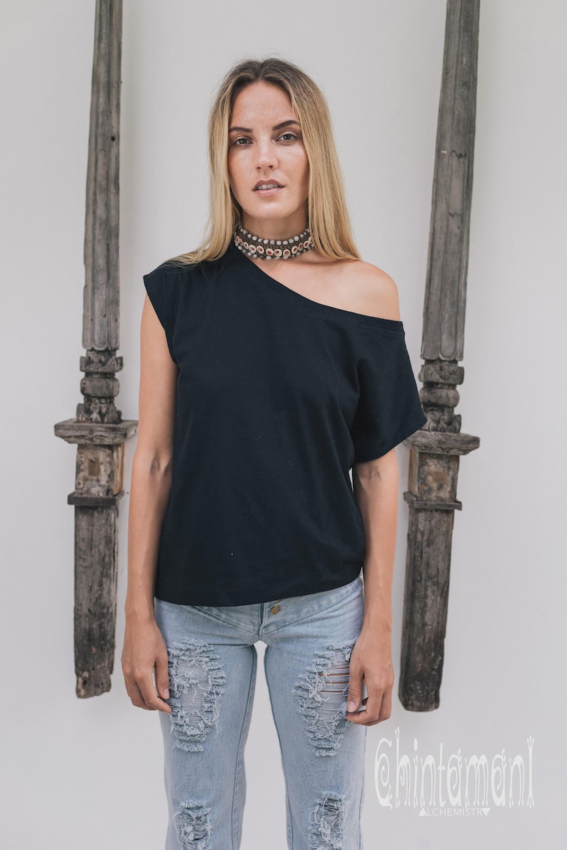 May include: Black off-the-shoulder top paired with light blue distressed jeans. The top has a single short sleeve and a unique asymmetrical neckline. A beaded choker necklace adds a touch of detail. The background features wooden decorative elements.