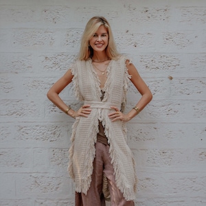 May include: A sleeveless, beige, open-front vest featuring a textured knit pattern, a waist tie, and long fringe detailing along the front edges and armholes. The garment is worn over a dark brown top and loose-fitting, light brown trousers.