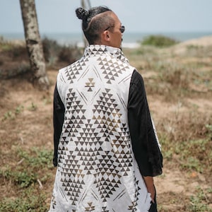 May include: A white vest with a geometric triangle pattern in black and gold is worn over a black long-sleeve shirt. The vest has a frayed hem. The image includes the text "chintamani alchymy".