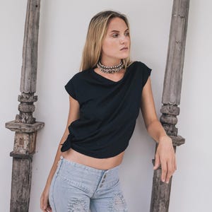 May include: A black t-shirt with a knotted side, paired with light blue distressed jeans. A silver beaded choker necklace is worn. The background includes weathered wooden posts and a white wall.