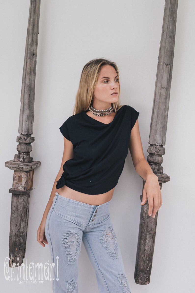 May include: A black t-shirt with a knotted side, paired with light blue distressed jeans. A silver beaded choker necklace is worn. The background includes weathered wooden posts and a white wall.