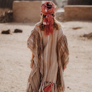Puede incluir: Un poncho a rayas beige y marr&oacute;n con largos flecos en los bordes, usado con un pa&ntilde;uelo de color coral con borlas colgantes. La prenda tiene una apariencia tejida y texturizada con patrones verticales en toda la tela.