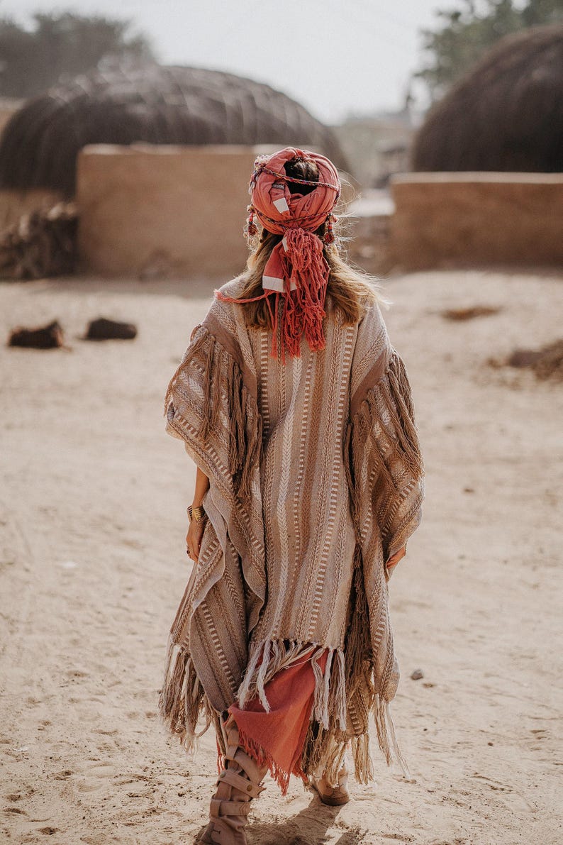 Puede incluir: Un poncho a rayas beige y marr&oacute;n con largos flecos en los bordes, usado con un pa&ntilde;uelo de color coral con borlas colgantes. La prenda tiene una apariencia tejida y texturizada con patrones verticales en toda la tela.