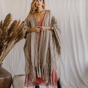 Puede incluir: Un kimono beige abierto por delante que presenta un patr&oacute;n tejido texturizado con rayas verticales y largos flecos a lo largo del dobladillo. La prenda se superpone sobre un vestido de color coral s&oacute;lido y se combina con botas de cuero negro.