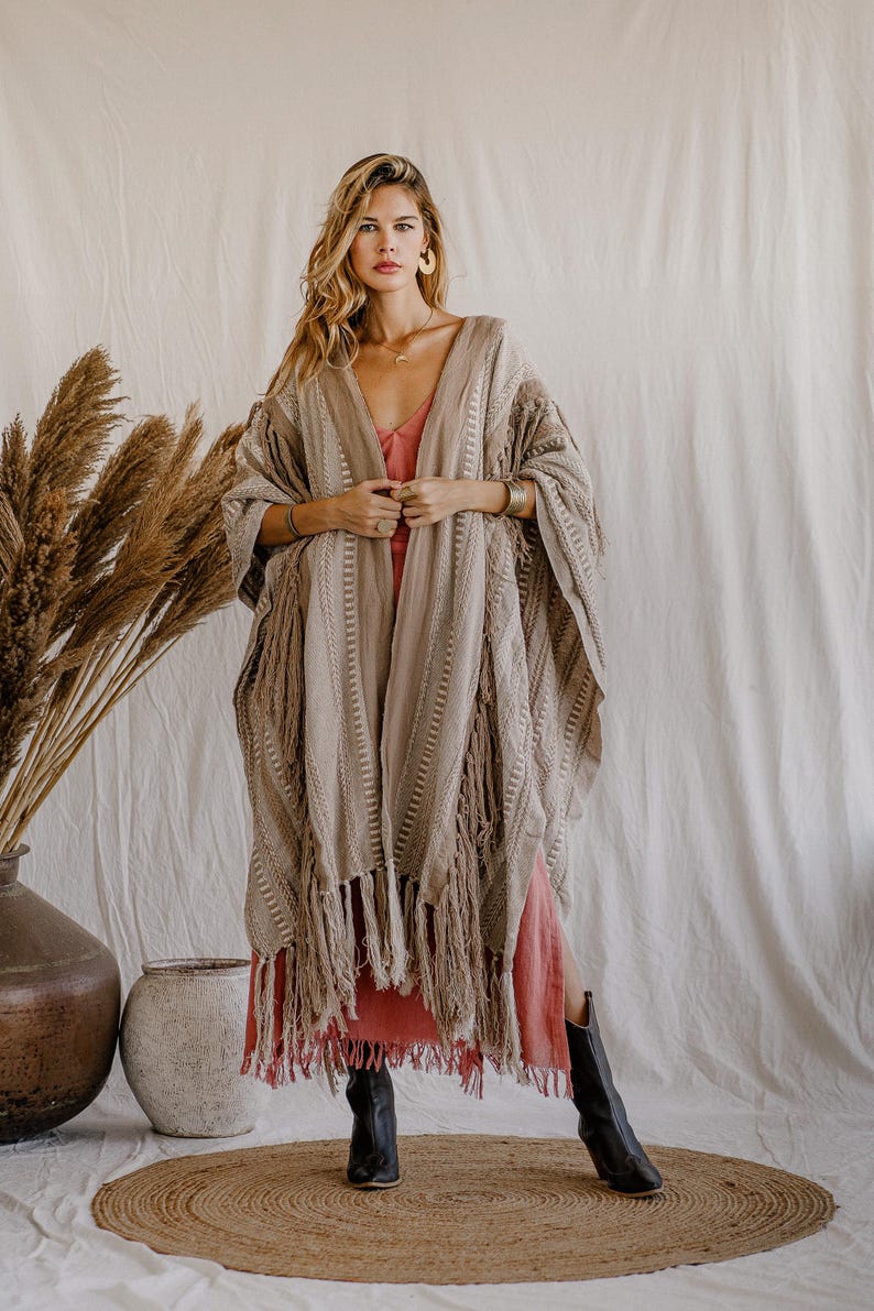 Puede incluir: Un kimono beige abierto por delante que presenta un patr&oacute;n tejido texturizado con rayas verticales y largos flecos a lo largo del dobladillo. La prenda se superpone sobre un vestido de color coral s&oacute;lido y se combina con botas de cuero negro.