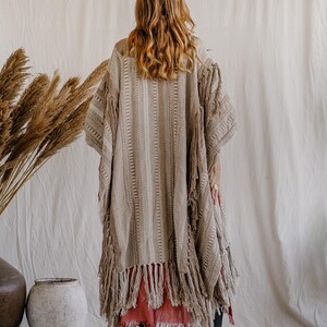 Puede incluir: Un kimono beige texturizado de frente abierto con patrones tejidos verticales y detalles de flecos largos a lo largo del dobladillo y las mangas. La prenda se superpone sobre una falda roja s&oacute;lida y se combina con botas de cuero negro.