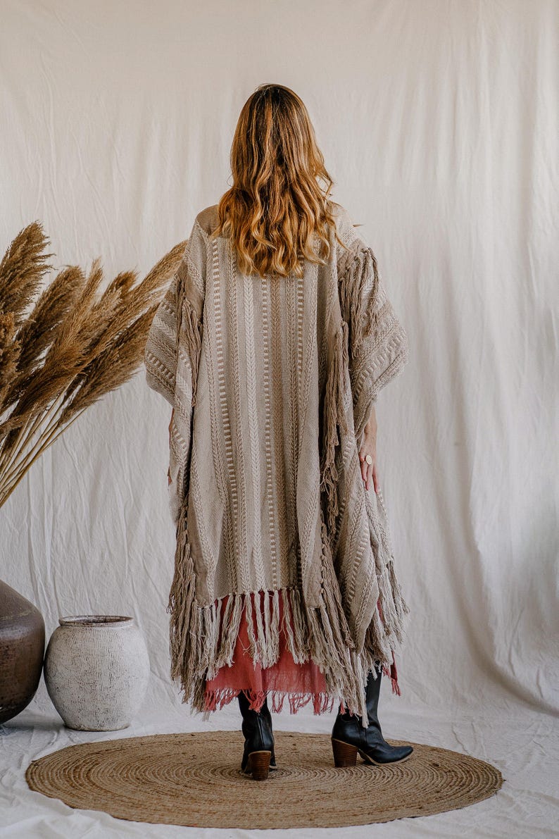 Puede incluir: Un kimono beige texturizado de frente abierto con patrones tejidos verticales y detalles de flecos largos a lo largo del dobladillo y las mangas. La prenda se superpone sobre una falda roja s&oacute;lida y se combina con botas de cuero negro.