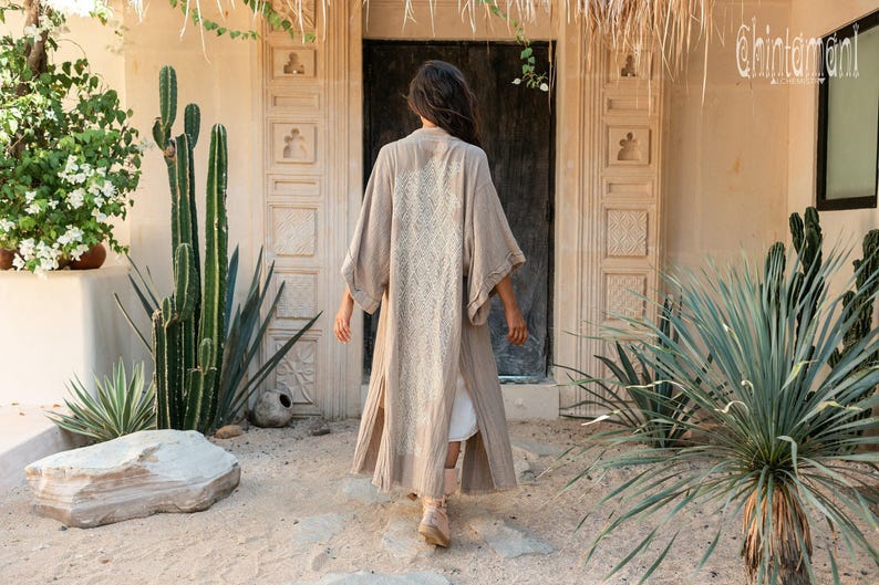 May include: A long, beige, open-front kimono-style robe featuring wide sleeves and a textured, cream-colored lace panel running down the center of the back. The garment is worn over a white base layer and reaches mid-calf length with side slits.