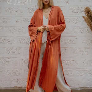 May include: A long, flowing rust-coloured robe with wide sleeves and a high slit, worn over a white top and linen trousers. The robe has a silky sheen and is paired with platform sandals. The word "chintamant" is printed on the upper left.