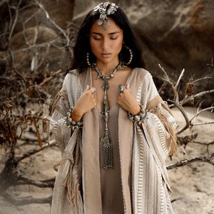 Puede incluir: Un kimono abierto a rayas beige y blancas con detalles de flecos, usado sobre un vestido beige liso. El conjunto se complementa con un tocado de monedas plateadas, pendientes de aro grandes, un collar largo con colgante y pulseras gruesas.
