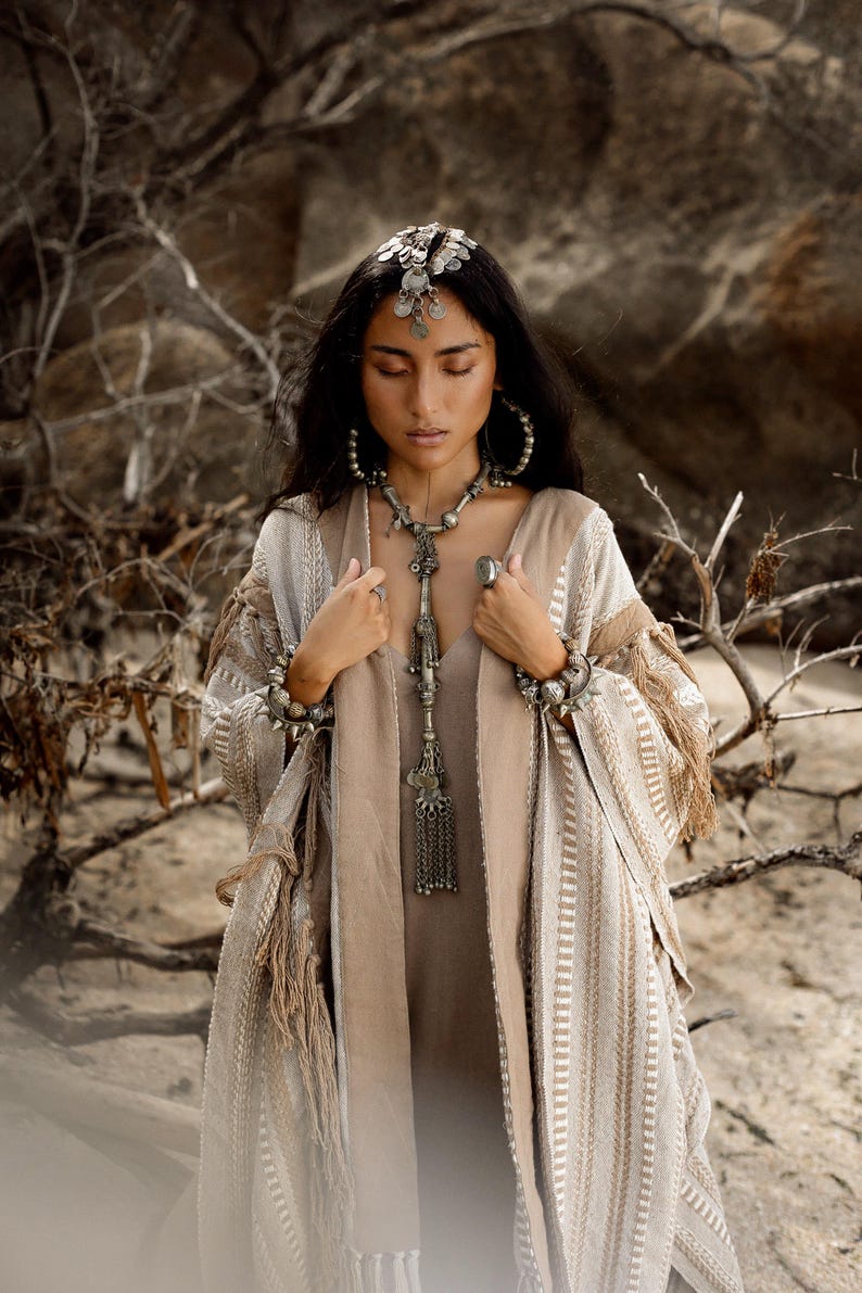 Puede incluir: Un kimono abierto a rayas beige y blancas con detalles de flecos, usado sobre un vestido beige liso. El conjunto se complementa con un tocado de monedas plateadas, pendientes de aro grandes, un collar largo con colgante y pulseras gruesas.