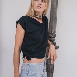 May include: Black draped-neck top with a knotted side detail. Paired with light-wash denim jeans. A beaded choker necklace adds a touch of detail. The image features a wooden decorative element and the text "chintamani alchemy".