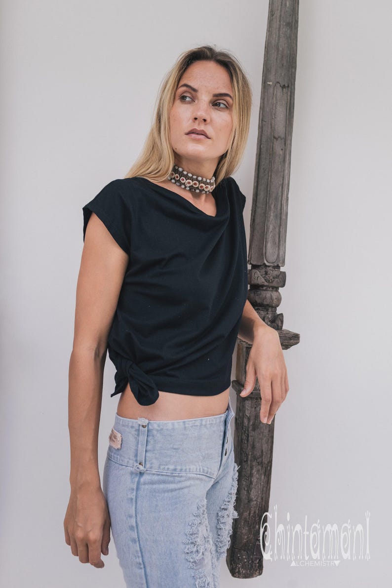 May include: Black draped-neck top with a knotted side detail. Paired with light-wash denim jeans. A beaded choker necklace adds a touch of detail. The image features a wooden decorative element and the text "chintamani alchemy".