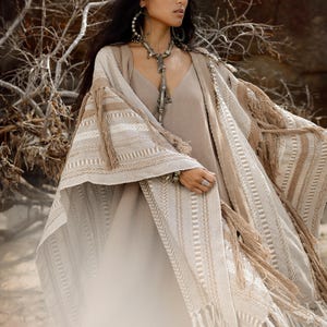Puede incluir: Un vestido beige sin mangas combinado con una capa texturizada abierta por delante, con rayas horizontales en tonos crema y tostado, rematada con largos flecos en los bordes. La prenda se complementa con un tocado y un collar de color plateado.