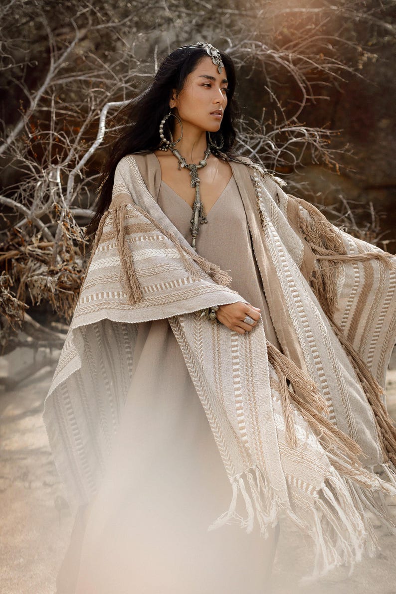Puede incluir: Un vestido beige sin mangas combinado con una capa texturizada abierta por delante, con rayas horizontales en tonos crema y tostado, rematada con largos flecos en los bordes. La prenda se complementa con un tocado y un collar de color plateado.